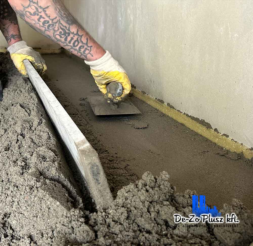 A képen egy szakember kézzel simítja az estrich betont friss bedolgozás után. A pontos kézi munka biztosítja a tökéletes padlóalapot, amely hosszú távon stabil és burkolásra kész. A De-Zo Plusz csapata az aljzatbetonozás minden lépésére figyelmet fordít.