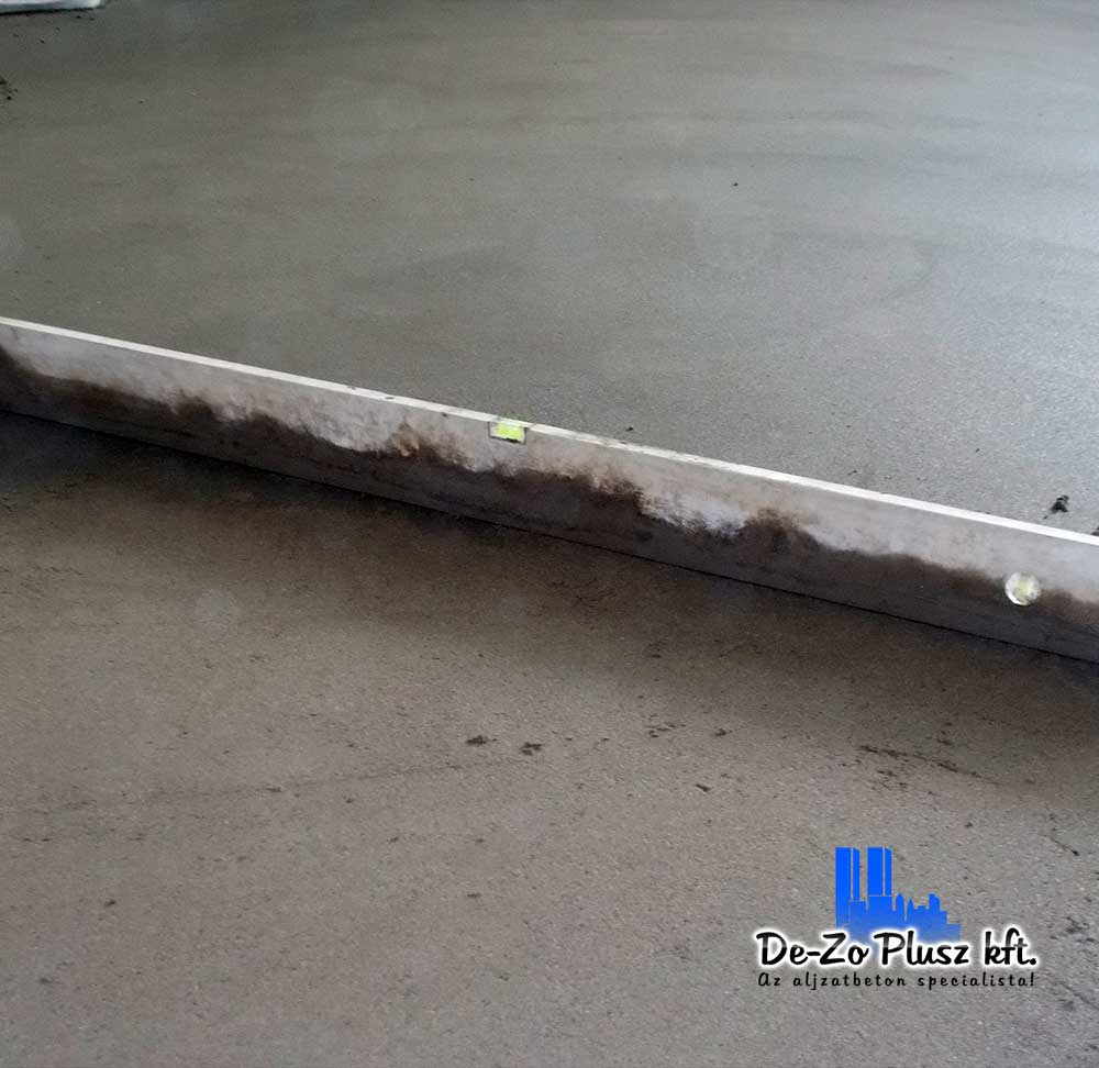 A képen frissen lehúzott estrich betonfelület látható egy épülő belső térben. A szintező léc éppen a beton simítási folyamatában van, jelezve az aljzatkiegyenlítés precizitását. Az egyenletes felület kulcsfontosságú a burkolatok hosszú távú stabilitása szempontjából. A kép hitelesen ábrázolja a kivitelezés egyik kritikus, mégis sokszor háttérbe szoruló lépését.